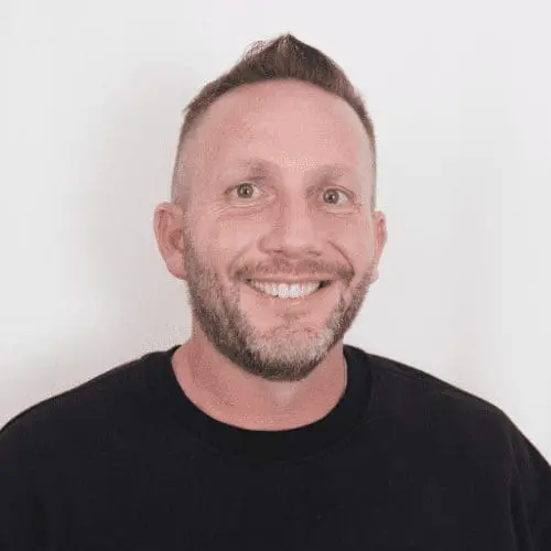 A smiling man with short hair and a trimmed beard, wearing a black crewneck sweater, stands in front of a plain white background, representing expertise in Salt Lake commercial construction and office buildout projects.