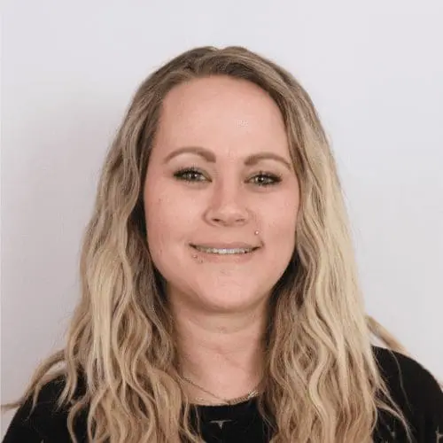 A woman with long, wavy blonde hair and light skin smiles at the camera. She is wearing a black top, with a light-colored background that resembles an office buildout or modern commercial remodel.