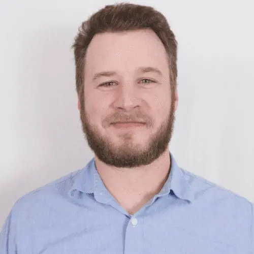 A man with short brown hair and a beard, wearing a light blue button-up shirt, smiles slightly while standing in front of a plain white background, ready to discuss your next office buildout or commercial remodel project.