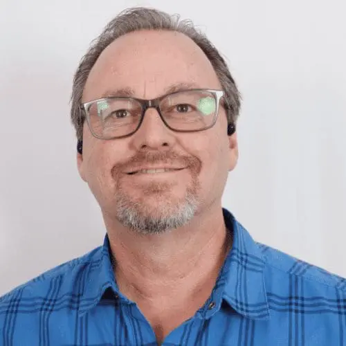 A middle-aged man with short brown hair, glasses, and a goatee smiles at the camera. He is wearing a blue plaid shirt and stands in front of a plain white background, ready to discuss your next commercial remodel project.