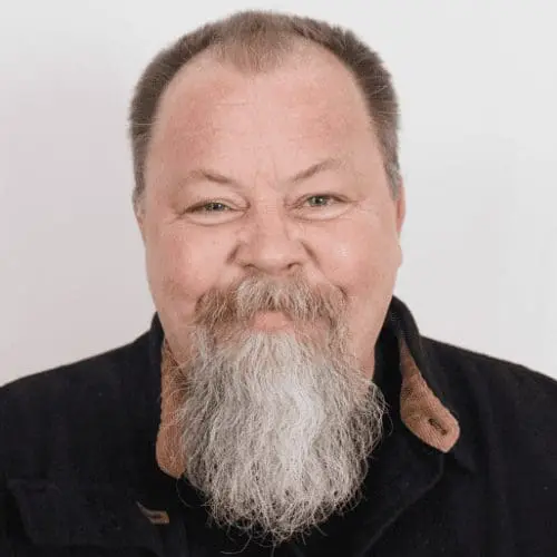 A middle-aged man with a long, gray beard and mustache smiles warmly. He has short, light brown hair and is wearing a dark jacket with a brown collar, ready for his next salt lake commercial construction project. Plain white background.