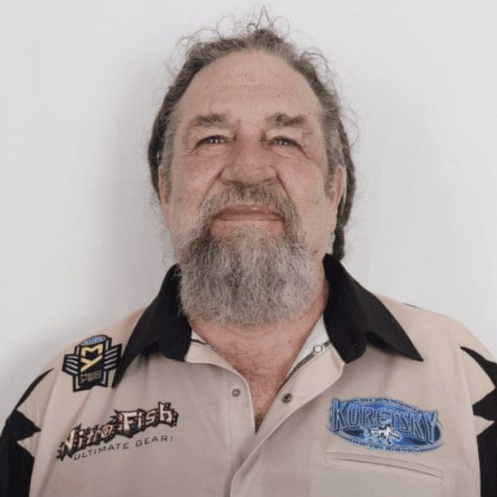 A man with long gray hair and a gray beard is wearing a tan and black collared shirt with embroidered patches that read Koretsky and Nitro Fish Ultimate Gear, standing in front of a plain white background, representing Salt Lake commercial construction.
