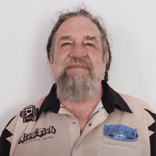 Bearded man wearing a beige shirt with logos, smiling against a plain background, representing the experienced team at AIS Commercial Construction.