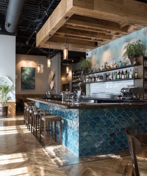 Modern restaurant bar featuring a blue tiled counter, wooden beams, and stylish lighting, with shelves displaying various beverages, surrounded by a welcoming interior design.